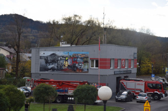 Mural na budynku Ochotniczej Straży Pożarnej. Widać dwie strony muralu - połowa jest monochromatyczna i przedstawia historycznych strażaków z cysterną wody ciągniętą przez konie. Kolorowa strona muralu to strona współczesna z wozem strażackim.