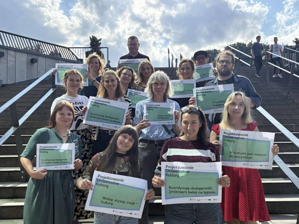 Zdjęcie grupowe z&nbsp;Laboratorium Dostępności. Kilkanaście osób uczestniczących w&nbsp;szkoleniu w&nbsp;Gdańsku stoi na&nbsp;schodach przed budynkiem muzeum. Każda osoba trzyma zieloną planszę z&nbsp;inspirującym hasłem dotyczącym dostępności w&nbsp;kulturze oraz nazwą projektu: „Projektowanie uniwersalne kultury”.