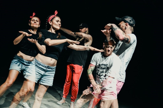 Na ciemnej scenie pięć osób uczestniczy w działaniach ruchowych i tanecznych. Uczestnicy stoją blisko siebie, wykonując różne gesty rękami i ramionami. Zdjęcie zostało wykonane z użyciem dłuższego czasu naświetlania, przez co część sylwetek i ruchów jest lekko rozmyta, co podkreśla dynamikę sceny. Osoby ubrane są w swobodne stroje sportowe w kolorach czarnym, czerwonym, białym i jasnoniebieskim.