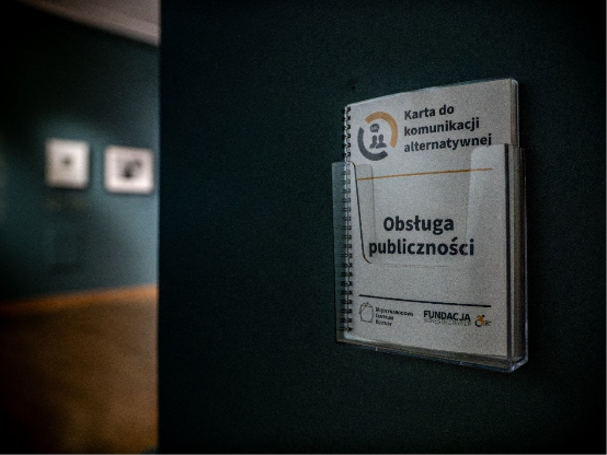 Karta do&nbsp;komunikacji alternatywnej dla obsługi publiczności umieszczona w&nbsp;przezroczystym uchwycie na&nbsp;ścianie galerii. W&nbsp;tle widoczne są rozmyte elementy ekspozycji muzealnej
