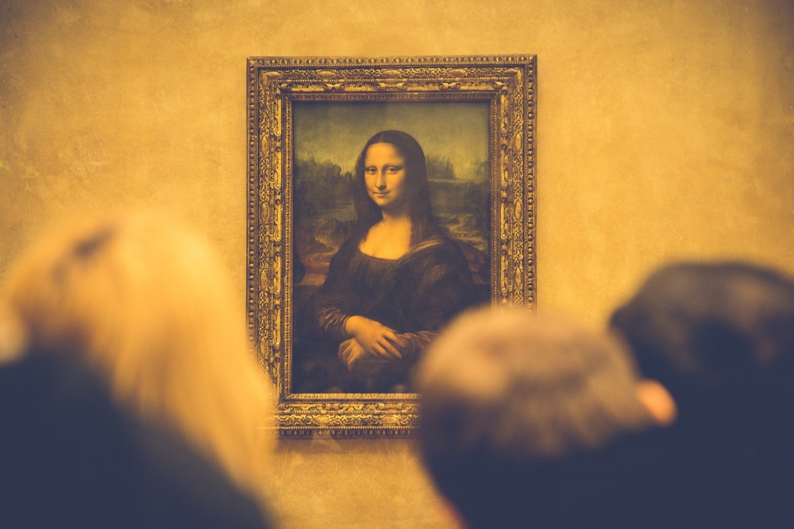 Na zdjęciu znajduje się portret Mona Lisy, w złotej ramie, wiszący na szarej ścianie. Na pierwszym planie widać tył głowy trzech osób, które patrzą na obraz. 