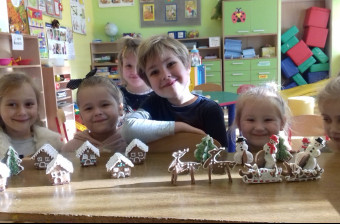 Uśmiechnięte dzieci siedzą przy stole, na którym znajdują się domki oraz sanie Świętego Mikołaja zbudowane z piernika.