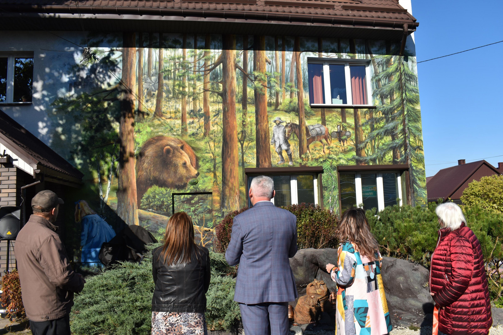 Ludzie stojący tyłem patrzący się na mural przedstawiający las a wnim niedźwiedzia oraz podróżnika prowadzącego dwa objuczone konie