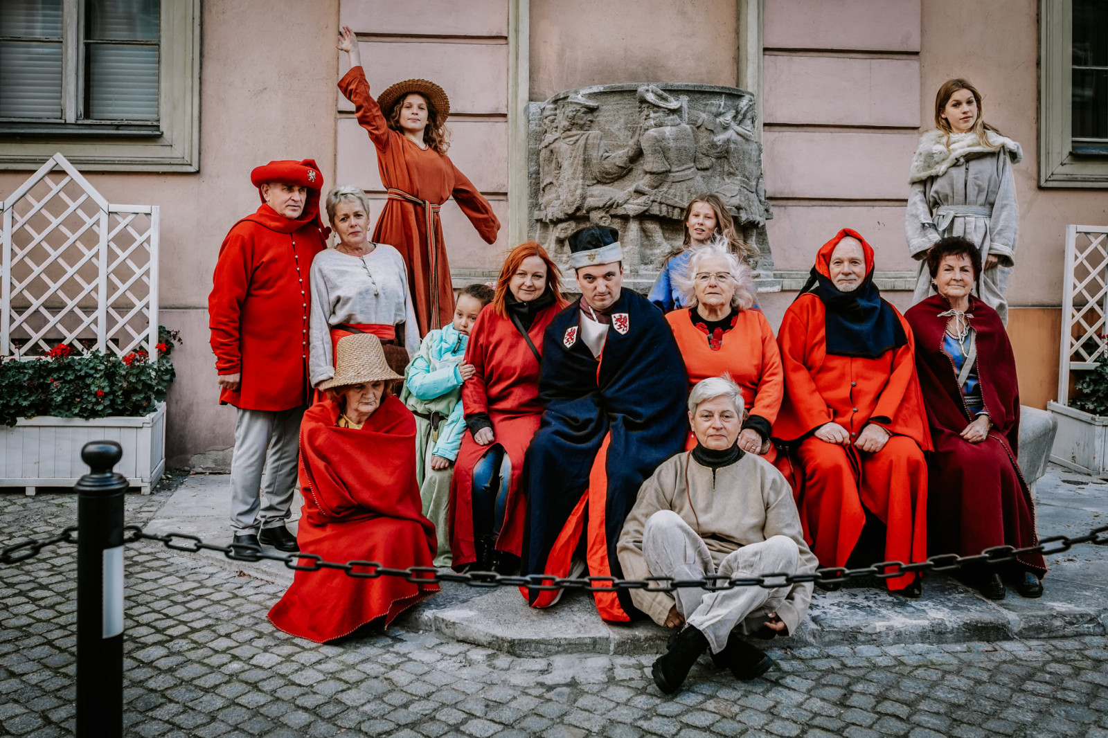 Trzynaścioro uśmiechniętych uczestników i uczestniczek warsztatów fotograficznych, w różnym wieku od dzieci po osoby starsze, pozują do zdjęcia siedząc na stopniach fontanny. Wszyscy są ubrani w stroje średniowiecznych mieszczan, które są w większości koloru czerwonego.