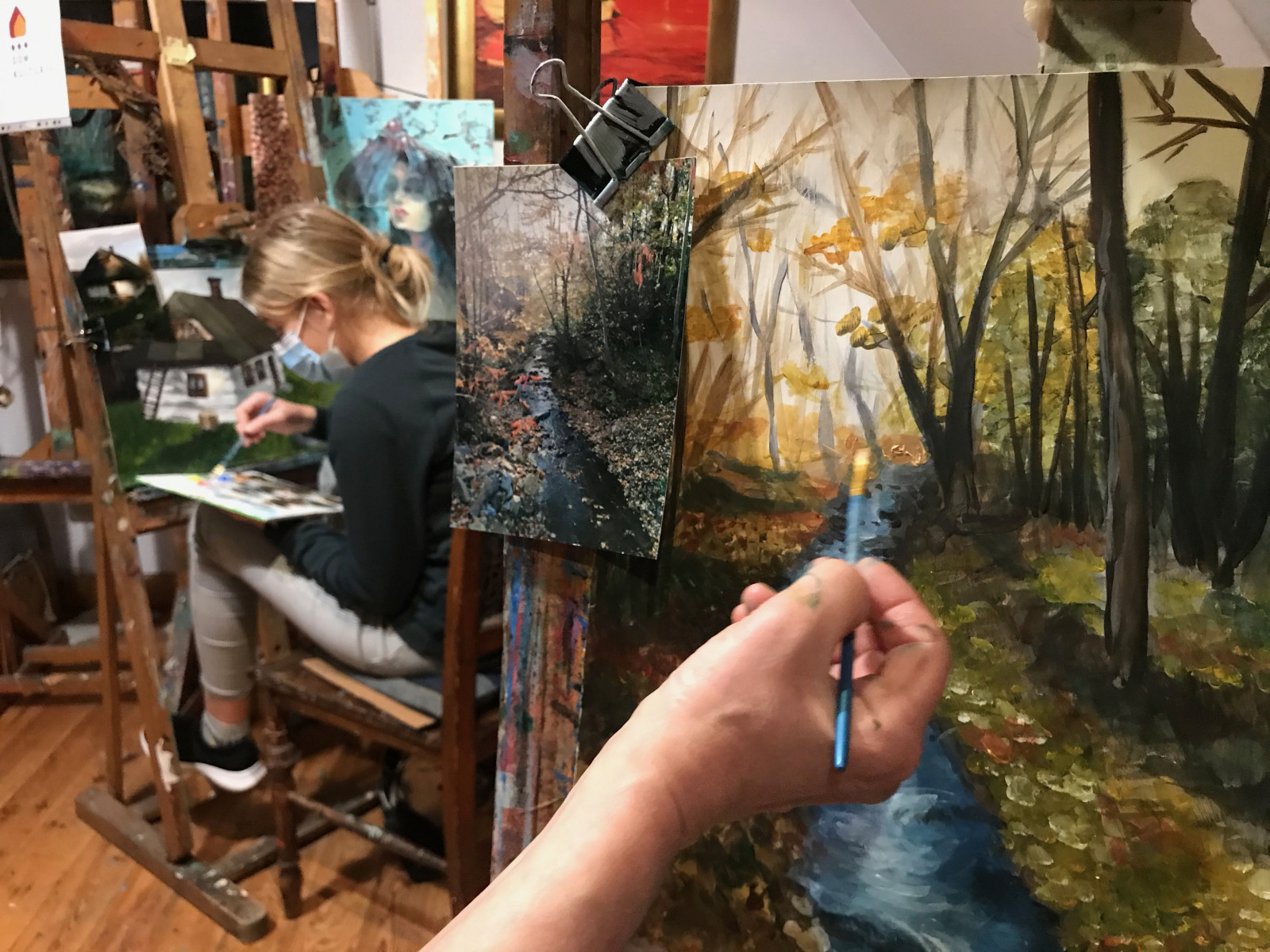 Na pierwszym planie sztaluga z namalowanym obrazem rzeki płynącej w lesie. Na drugim planie dziewczynka przy sztaludze maluje stary dom.