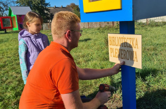 Mężczyzna instaluje drewnianą, kolorową budkę z żółtymi drzwiczkami. Obok niego stoi dziecko.